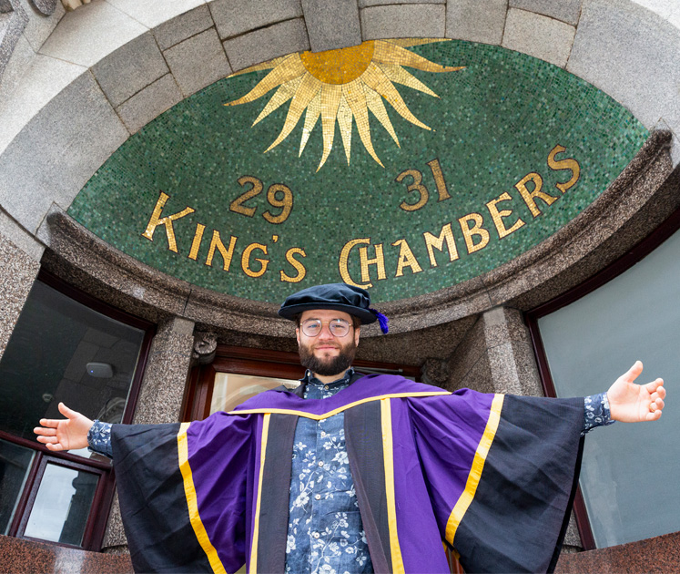 A male PhD student stands hands outspread under the mosaic entrance of the King's Chambers on campus
